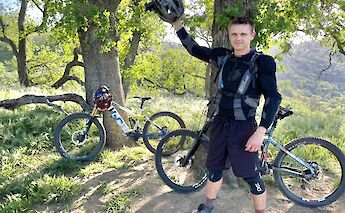 A man in biking gear is standing next to two mountain bikes under trees, holding a helmet in the air. The scene is set in a sunny, wooded area.