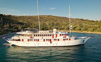 A white yacht named Bellezza with multiple decks is anchored near a tree-lined coastline, set…