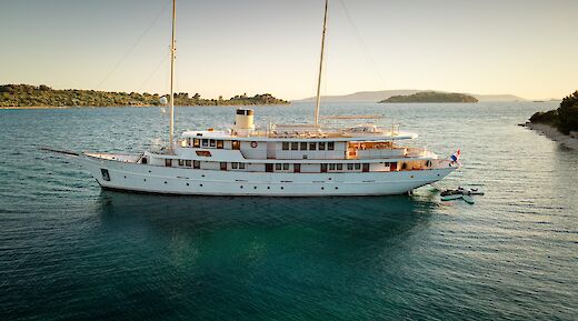 A white yacht named Bellezza with two masts is anchored in a calm body of water, surrounded by small islands and a distant coastline under a clear sky.