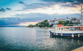 A coastal view of Skiathos, Greece, with a boat docked in the foreground and a hillside dotted with white buildings in the background.