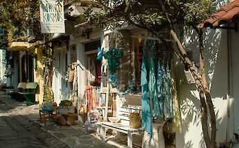 A narrow street in Skiathos Town, Greece, lined with small shops displaying colorful clothing and accessories.