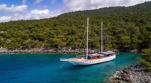 A sailboat named Milo M is anchored in a secluded cove with clear turquoise water, surrounded by lush green hills and rocky shores under a partly cloudy sky.