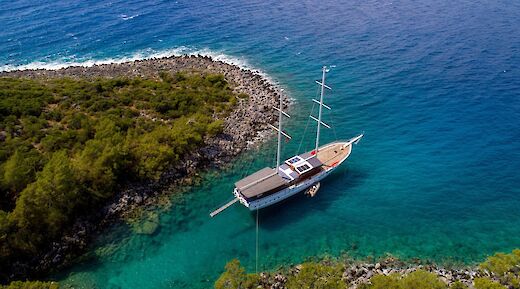 Aerial view of the Milo M, a boat anchored in turquoise waters near a rocky coast with dense green vegetation.
