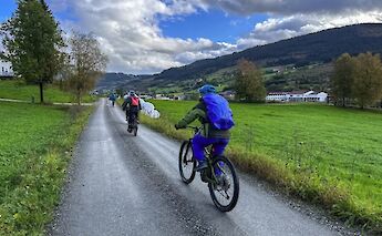 Cyclists riding e-mountain bikes on a country road flanked by green fields and hills in Voss, Norway.