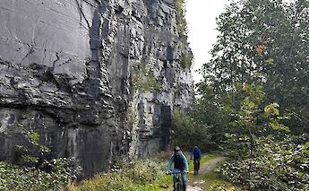 Cyclists on a trail under a sheer rock face with lush greenery in Voss, Norway.