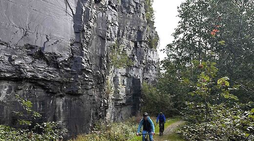 Cyclists on a trail under a sheer rock face with lush greenery in Voss, Norway.