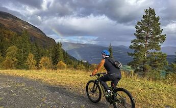 A person riding an e-mountain bike on a trail in Voss, Norway, with a rainbow in the sky above a valley surrounded by trees and mountains.
