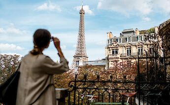 A person gazing at the Eiffel Tower from a Parisian balcony, framed by autumn trees and classic architecture.