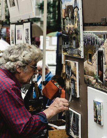 An artist working on paintings displayed at an outdoor art market in Paris, France, with various artworks depicting architectural scenes.