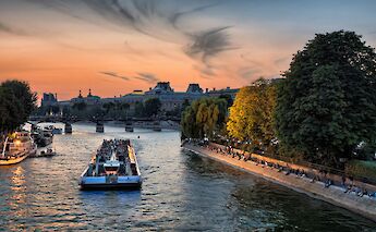 A sightseeing boat on the River Seine at sunset in Paris, France, with people relaxing along the riverbank and a colorful sky.
