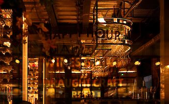 The warmly lit interior of a bar in Paris, France, featuring hanging wine glasses and bottles with a reflective window showcasing the cozy atmosphere.