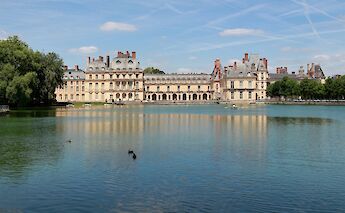 A large lake reflecting the facade of Fontainebleau Castle in France, surrounded by trees and under a clear sky.