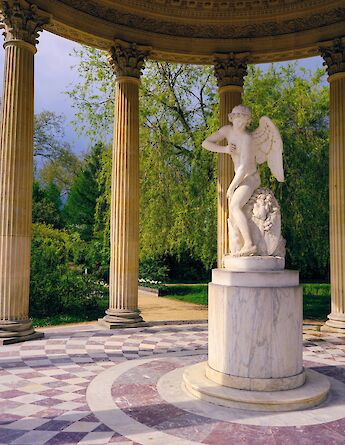 A marble statue of a winged figure inside a classical pavilion with columns in the gardens of Versailles, France.