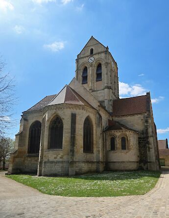 Notre-Dame-de-l'Assomption Church, a historic stone structure with a clock tower, stands against a clear blue sky in Auvers-sur-Oise, France.