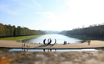 A sculpture situated by a canal in the gardens of Versailles, France, with expansive lawns and trees lining the waterway.