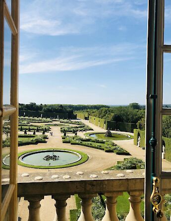 A view of the formal gardens of Versailles, featuring fountains and manicured hedges, seen through an open window.