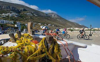 Cyclists ride along a coastal path on a sunny day, with a view of mountainous terrain and white buildings in the background.