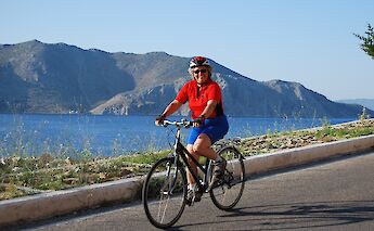 A person cycling along a coastal road with blue water and mountainous terrain in the background, in Greece.
