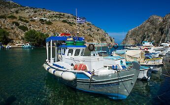 Fishing boats in a small harbor with a Greek flag, surrounded by rocky hills and clear blue water. Greece.
