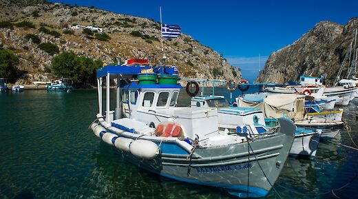 Fishing boats in a small harbor with a Greek flag, surrounded by rocky hills and clear blue water. Greece.