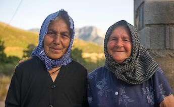 Two women wearing headscarves stand outdoors with a mountainous landscape in the background.