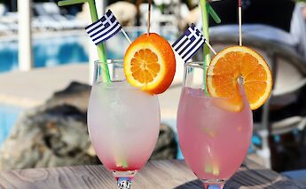 Two cocktails with orange slices and umbrellas by a swimming pool, featuring Greek flags.