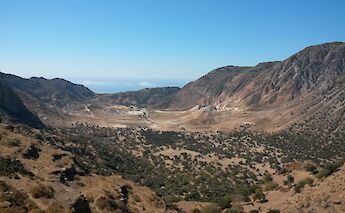 A volcanic crater nestled between rugged hills on Nisyros Island, Greece, with sparse vegetation and the sea visible in the distance.