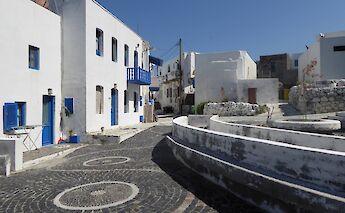 A cobblestone path winds through a village with white buildings and blue accents in Mandraki on Nisyros Island, Greece, under a clear blue sky.