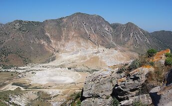 The caldera of the Nisyros volcano in Greece, showing a rugged, barren landscape with rocky slopes under a clear blue sky.