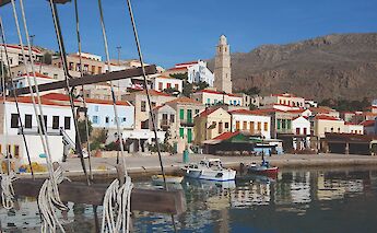 Colorful buildings with red-tiled roofs line the waterfront in Symi, Greece, reflecting in the calm harbor water.