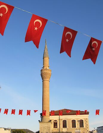 A mosque with a tall minaret, surrounded by Turkish flags with a crescent and star, under a clear blue sky.