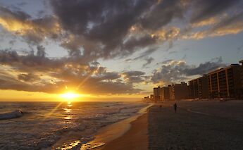 Sunset view at the Gulf of Gökova, Turkey, with waves breaking on a sandy beach and buildings in the distance.