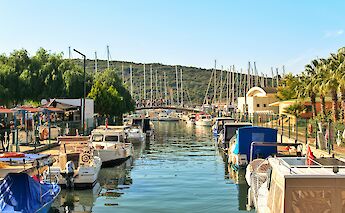 A marina in Bodrum, Turkey, with boats docked along the canal and a pedestrian bridge in the background.
