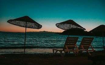 Sunset view of sun loungers and straw umbrellas on a beach in Bodrum, Turkey, with the sea and mountains in the background.