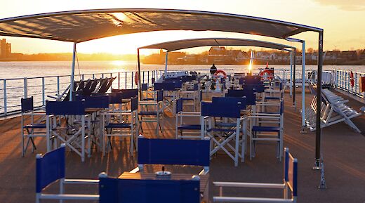 The sun deck of the "Lisabelle" boat at sunset, featuring arranged tables and chairs under a canopy, with a view of the water.