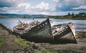 Two old and weathered fishing boats beached on the shore of the Isle of Mull, Scotland, with a scenic view of the water and hills in the background.