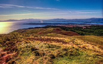 A scenic view overlooking the hilly landscape around Oban, Scotland, with a distant view of the sea and mountains under a clear sky.