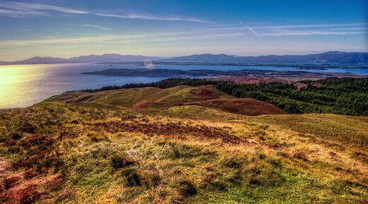 A scenic view overlooking the hilly landscape around Oban, Scotland, with a distant view of the sea and mountains under a clear sky.