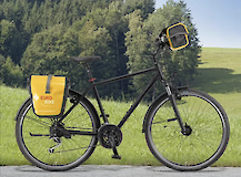 A black touring bike with a yellow saddlebag and handlebar bag, placed against a backdrop of green grass and trees.